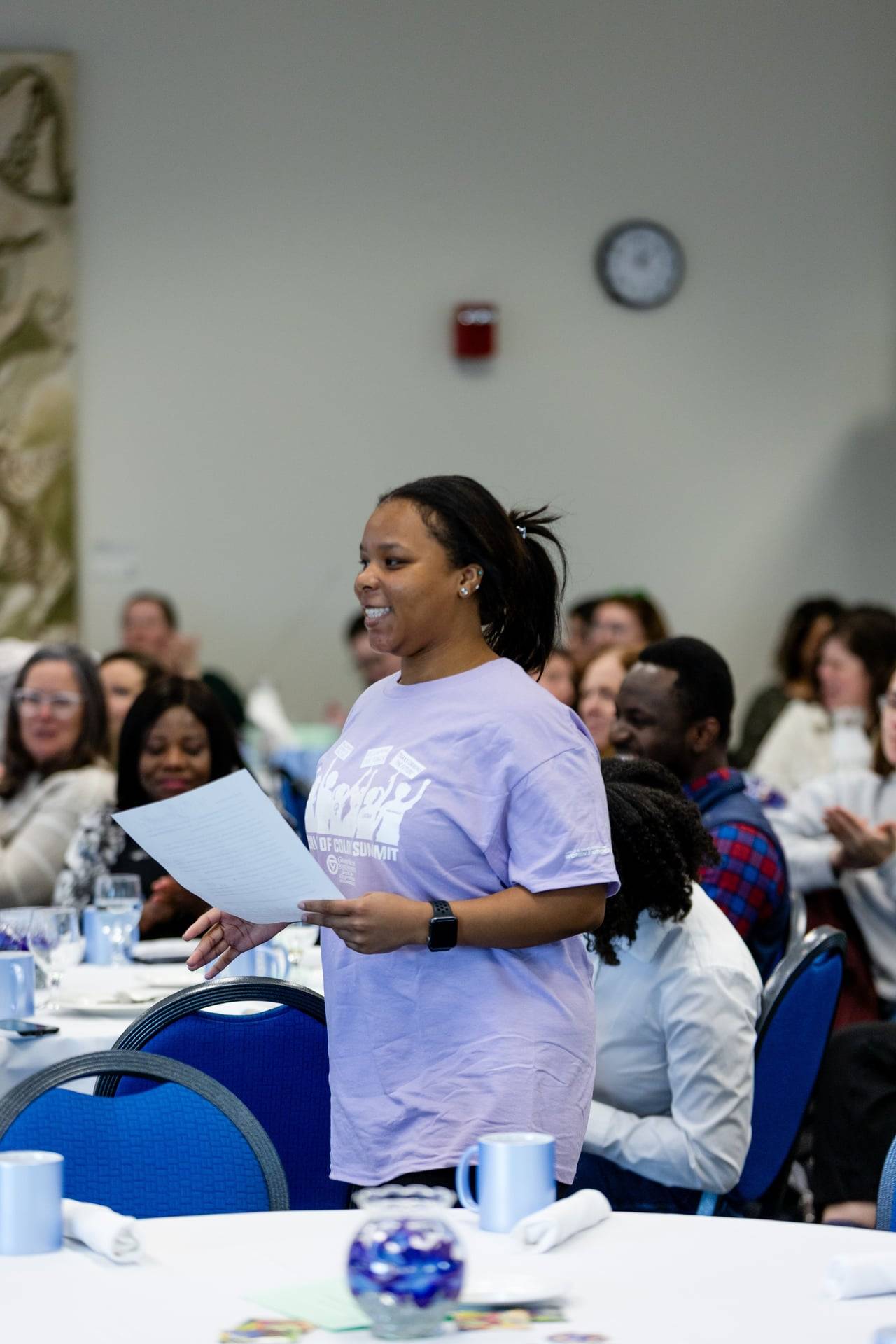 Photo shows a student in a lavender T-shirt smiling and holding a piece of paper while members of a seated crowd look on, smiling
